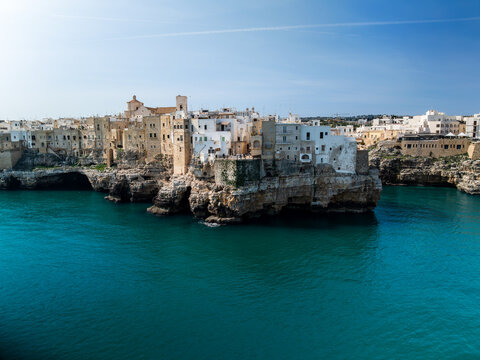 Aerial view of historic white buildings perched on rugged limestone cliffs above the turquoise Adriatic Sea under a clear blue sky in Polignano a Mare, Apulia, Italy.