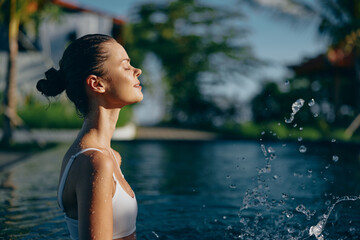 Fototapeta premium A woman at a sunlit poolside, eyes closed, enjoying a breezy outdoor moment as water sprays gently, conveying relaxed lifestyle and summer calm amid lush greenery and bright natural colors