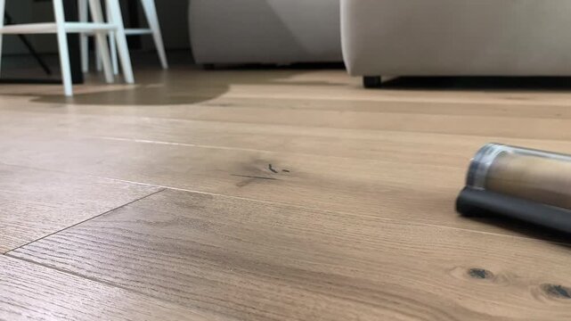 Vacuum cleaner removes dust from wooden floor using rotating brush. Close shot shows surface cleaning with modern device. Home routine maintains hygiene and neat organized interior..