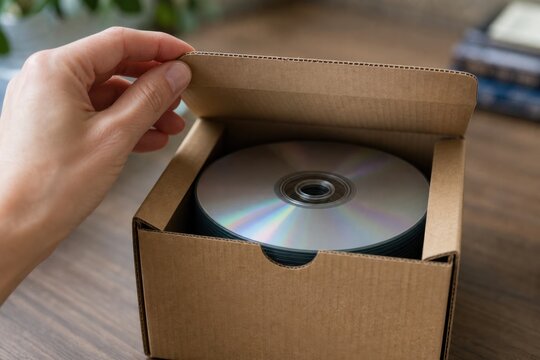 Hand opening a cardboard box with CDs inside to reveal a stack of shiny compact discs or DVDs being delivered or organized for media storage and collection
