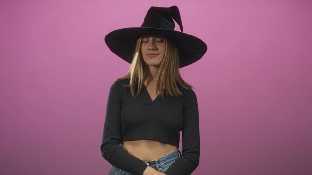 Woman wearing a wide black witch hat, adjusting her hands over exposed midriff and smiling while standing against a pink studio backdrop; playful.