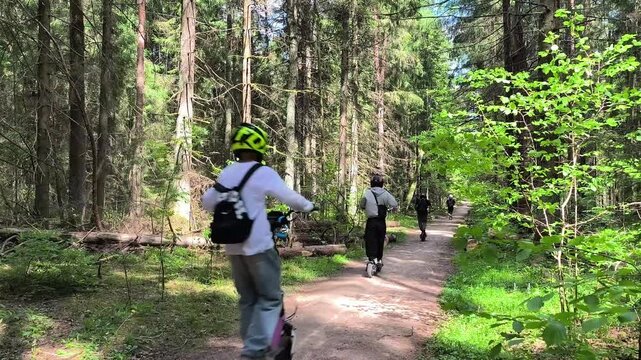 People ride electric scooters along a forest trail. The group moves through a green woodland path in sunlight. Outdoor recreation and eco transport concept..