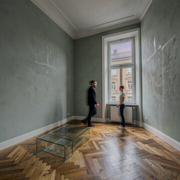 Two men walking in a spacious room with green walls and wooden floor near window.