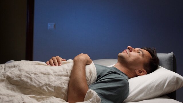 Man lying awake in bed at night, staring at the ceiling, struggling with insomnia and racing thoughts in a dark bedroom, tense and restless in quiet solitude