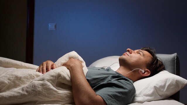 Man lying on a comfortable bed in a dark bedroom, listening to an audio podcast or meditation through wired earphones, finding peace and relaxation during his evening routine