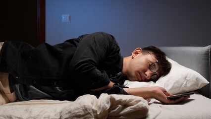 Fototapeta premium Man lying on his side in bed while sleeping, still holding his smartphone, exhausted from late night use and possibly experiencing digital eye strain, making it difficult to achieve restful sleep