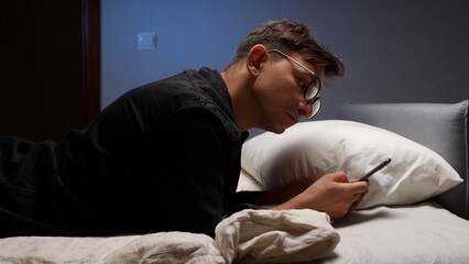 Obraz premium Young man wearing glasses lying in bed, scrolling on a mobile phone in a dark room, displaying internet addiction and the negative impact of blue light on sleep before bedtime