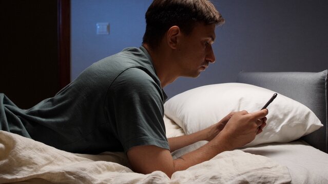 Man lying in bed at night scrolling smartphone, blue screen light on his face, sleepless and scrolling in dark bedroom, modern tech addiction and disrupted sleep routine