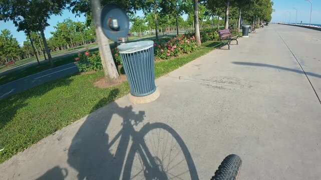 Bicycle POV. A shadow silhouette moving along the concrete pathway, past blooming rose flower beds and green trees in Orhangazi City Park, Maltepe
