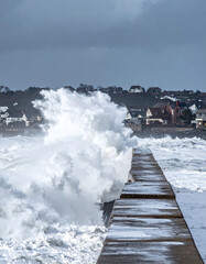 Obraz premium A powerful stormy ocean wave crashes spectacularly against a concrete pier, sending massive white sea spray high into the air against a coastal town backdrop under dark winter skies.