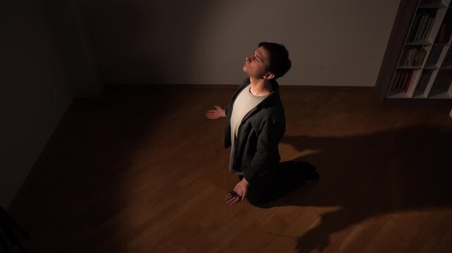 Man kneeling on a wooden floor with hands raised toward a single beam of light, seeking spiritual solace, prayerful reflection and inner peace in a quiet, dark room