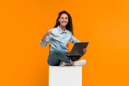 A woman sits on a box while using her laptop. She wears a light blue shirt and jeans. She smiles and shows a thumbs up against a bright orange background