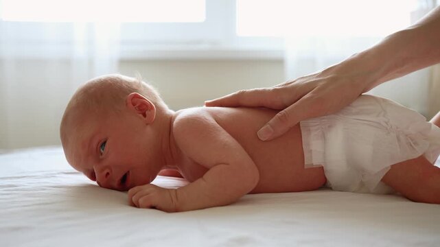 Newborn baby in a diaper is lying on its stomach for tummy time, crying while trying to lift its head as a parent's gentle hand provides comfort by caressing its back