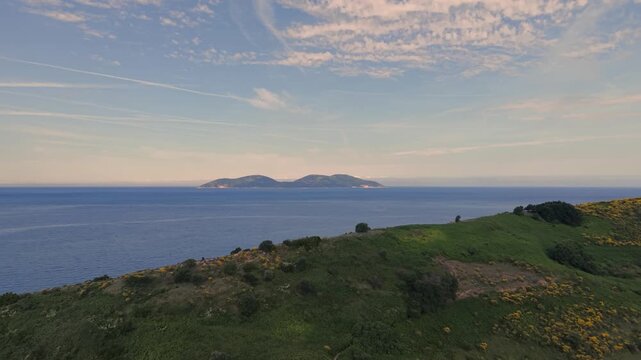 Aerial view of Sazan Island across the blue sea from the lush green hills covered in yellow wildflowers under a soft sky in Vlore, Vlore County, Albania.