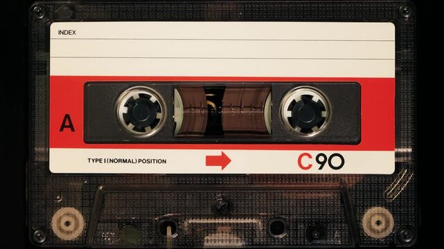 Close up view of generic retro audio cassette tape playing forward on side A showing red arrow and C90 index label against black background