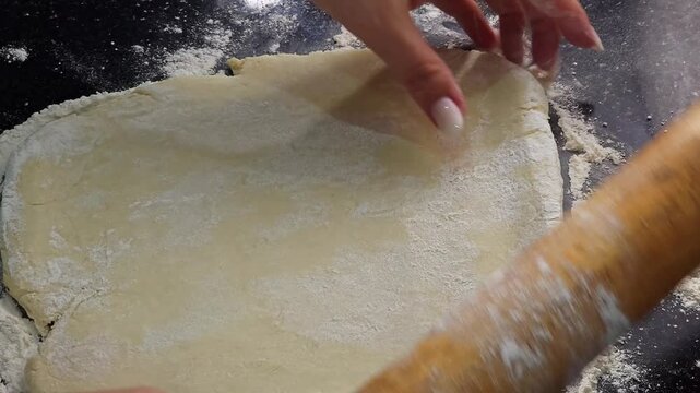 Top view close up of female hands rolling out fresh shortcrust pastry dough with a wooden rolling pin on a dark flour-dusted surface. Homemade baking process, cooking preparation concept.