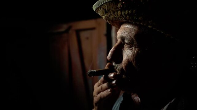 An older man in a straw hat pauses in quiet reflection while cigar smoke swirls through the night air. Selective lighting highlights facial features and creates a powerful mood of experience.