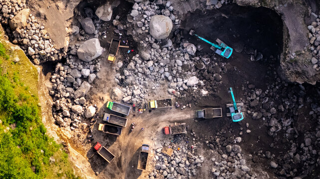 Aerial view of a stone quarry with excavators and dump trucks working among large boulders and workers on the rocky ground in Indonesia.