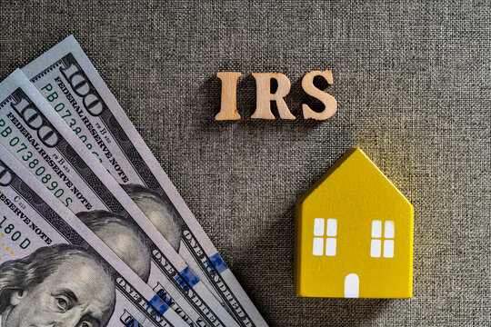 Property tax concept with IRS text wooden house and dollar bills. Top view wooden IRS letters with miniature yellow house and US dollar banknotes on grey background