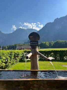 Wooden water fountain surrounded by lush greenery, with the Alps visible in the background. Scenic mountain landscape in Bavaria, Germany.
