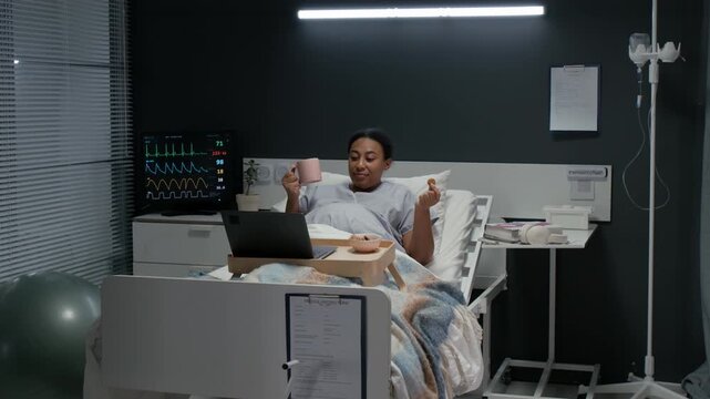 Black female patient lying in hospital bed,eating snack and drinking from mug while watching laptop. Video is suitable for healthcare and patient recovery campaigns, wide shot
