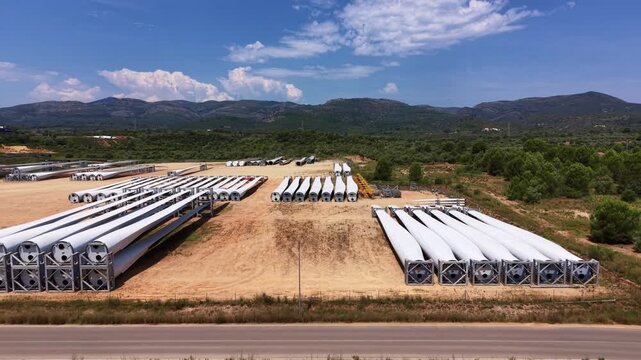 Rows of large white fiberglass wind turbine rotor blades stored on steel transport frames in an outdoor storage yard with green mountains and blue sky in background