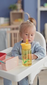 Toddler blonde girl drinking from yellow sippy bottle on a white highchair tray in a home dining corner inside building; quiet curiosity.
