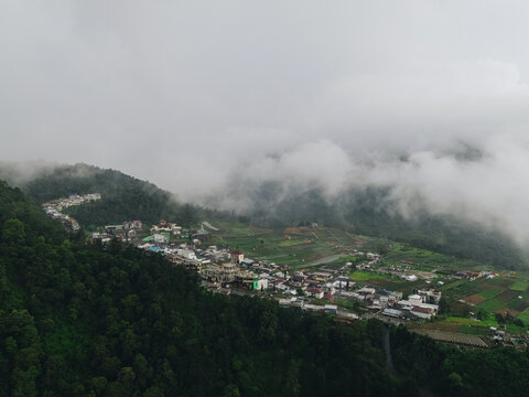 Aerial view of a mountain village nestled among lush green forests and agricultural fields under thick rolling mist in Tawangmangu, Central Java, Indonesia.