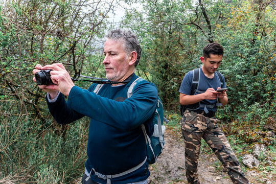 Father photographing nature with a camera while son uses a smartphone, showing generational difference in technology during outdoor activity