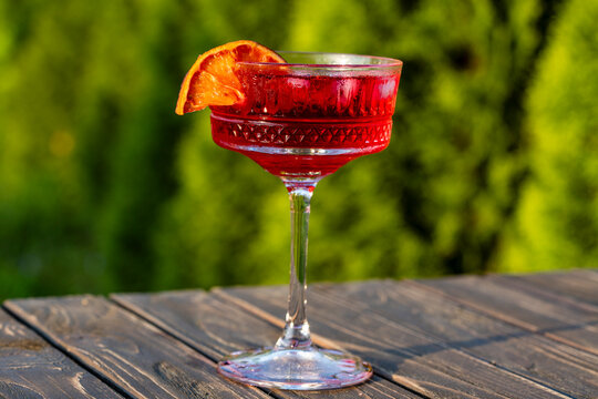 Blood orange margarita cocktail in coupe glass on wooden table. Refreshing red negroni or campari cocktail with ice cubes and orange garnish in a glass on a sunny day