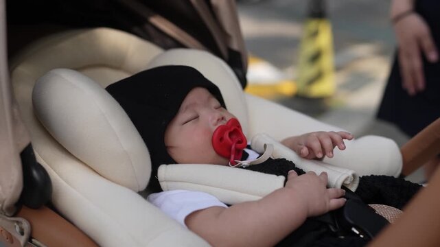 Tiny baby sleeps peacefully in car seat. Head covered with soft black hat. Red pacifier rests in mouth. Baby's hands rest gently on chest. Calm and relaxed, baby rests in white car seat