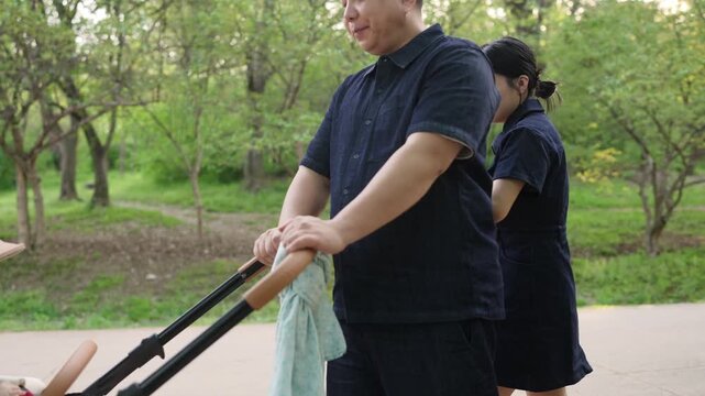 Couple walks through a park with a baby carriage. The man pushes the carriage gently. The woman holds a towel, watching nearby. They appear calm and content. Peaceful family outing