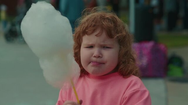 Ginger kid eating white cotton candy. Curly-haired child enjoys sugary snack in public park. Summertime treat for toddler experience, sweet dessert fun concept.