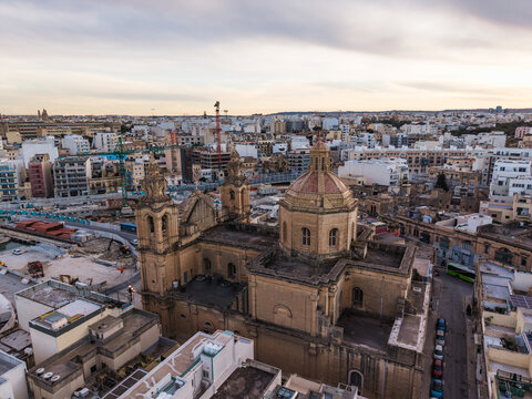 Aerial view of St. Joseph's Parish Church with its stone dome and bell towers overlooking urban rooftops and construction sites in Msida, Eastern Region, Malta.