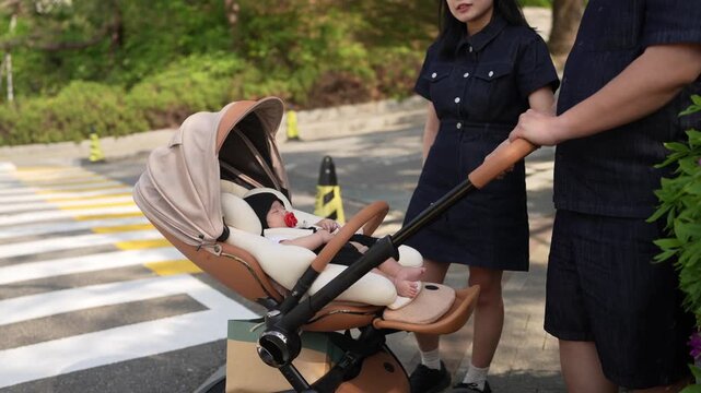 Baby rests peacefully in a stroller. Two adults stand nearby, quietly observing. The stroller sits at a crosswalk, surrounded by greenery. The baby appears calm and asleep. The scene suggests a quiet