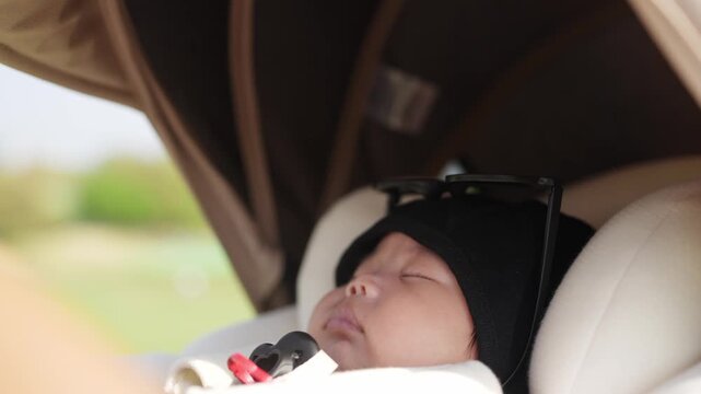 Baby sleeps peacefully in stroller seat. Soft light illuminates calm, peaceful face. Tiny hands rest gently against skin. Outdoor background blurred, natural green visible. Ideal for parenting, family