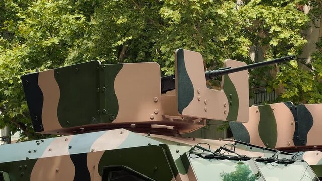 A close up shot of a camouflage armored military vehicle turret and machine gun, parked on a city street with lush green trees visible in the sunny background.

