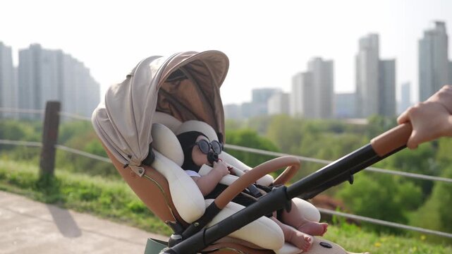 Soft light glows over city skyline. Baby sleeps peacefully in stroller. Gentle hands guide the pushcart forward. Urban green path winds beside tall buildings