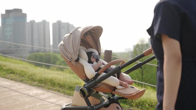 Soft sunlight glows over urban park paths. Stroller carries a calm baby wrapped in gentle tones. Parent's hand guides the push bar with care. Modern buildings loom softly in the background