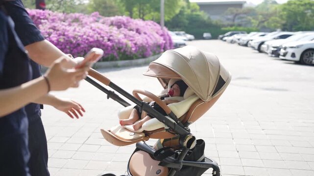 Soft sun glows on a baby's face. Baby sleeps peacefully in a stroller. Mother holds phone, captures moment. Purple flowers bloom behind the stroller. Parking lot stretches into the distance