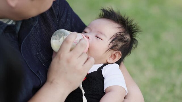 Soft grass backdrop frames tender moment. Baby with spiky hair drinks calmly from bottle. Parent's hands cradle child with care. Warm, peaceful scene of nurturing care. Ideal for parenting, child care