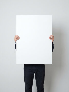 Unrecognizable person holding a large blank white poster board in front of face. Empty placard mockup with copy space for advertising, presentation, or message against a grey background