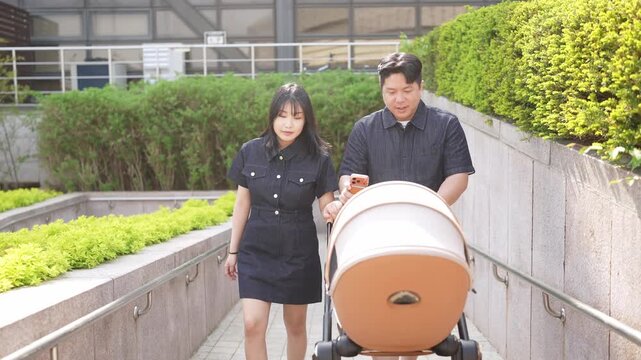 Young couple walks with stroller along paved path. They move together, smiling gently at each other. Green hedges line the walkway beside concrete walls. Natural daylight highlights their calm