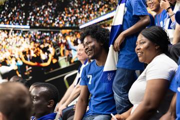 Fototapeta premium Diverse blue team football supporters chanting in packed stadium stands during soccer match, fan scarves and excited crowd atmosphere. Father, mother and son sharing family fun with laughter.