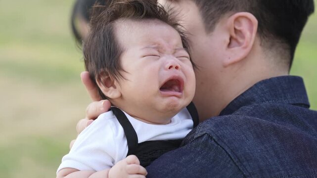 Tiny baby cries loudly, eyes squeezed shut. Mother's face shows soft concern beside father. Father holds child close, offering comfort. Outdoor setting gives peaceful, natural feel