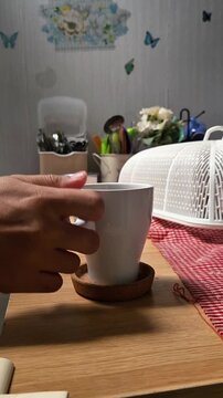 Person picking up a white coffee cup from a dining table for a morning sip, side view with warm filtering sunlight.