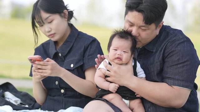 Young parents sit together, holding baby. Mother looks at phone, ignoring crying infant. Father comforts baby, eyes filled with tenderness. Natural daylight, outdoor park setting