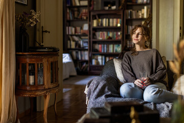 Young woman comfortably relaxing on a patterned sofa in a warm living room, holding a glass while looking away, surrounded by a bookshelf filled with books and a vintage bar cabinet © Nasos Zovoilis