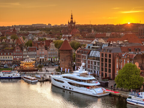 Aerial view of a luxury yacht docked at the Motlawa river waterfront with historic old town architecture and a carousel during sunset, Gdansk, Pomeranian Voivodeship, Poland.