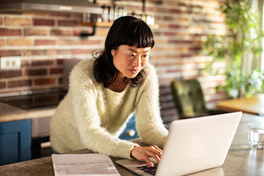Focused woman using laptop in home kitchen with paperwork